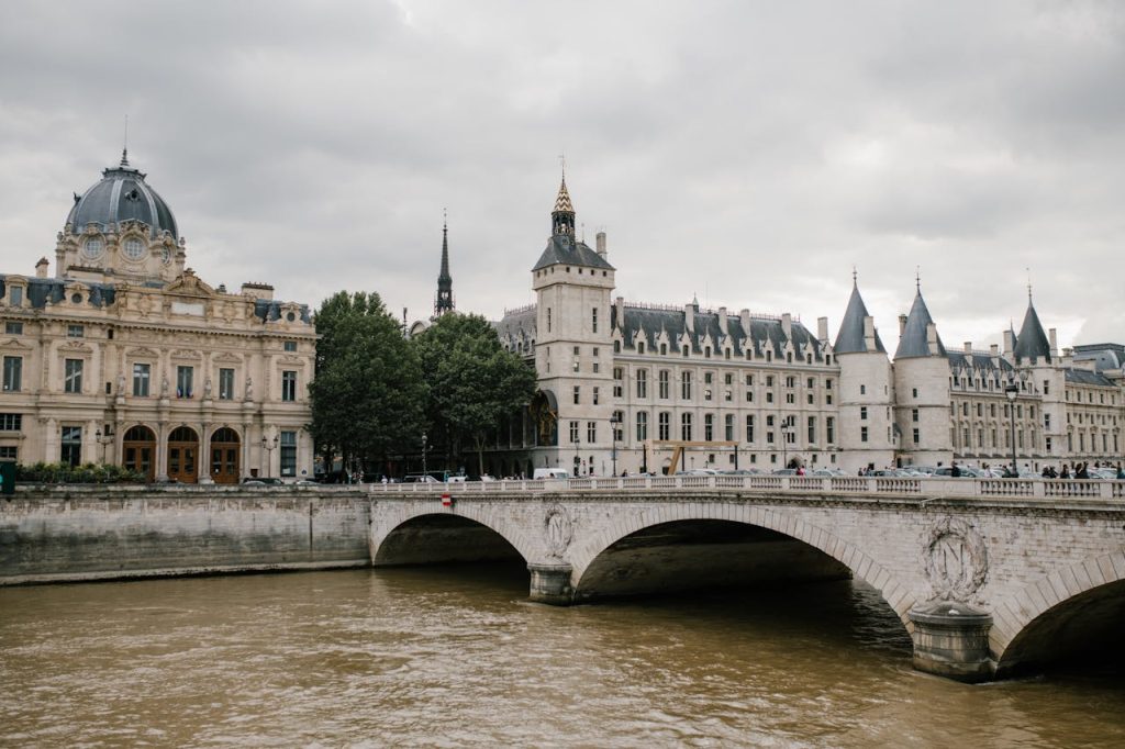 Aged stone bridge crossing river with architectural monument Conciergerie castle located in Paris under cloudy sky